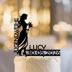 Könnte beinhalten: Eine zweistöckige weiße Torte mit dekorativem Zuckerguss. Ein schwarzer Tortenaufsatz zeigt die Silhouette einer Person, die eine Blume hält, mit dem Text "JUGENDWEIHE" und "LUCY 30.05.2026". Der Hintergrund ist mit Bokeh-Lichtern verschwommen.