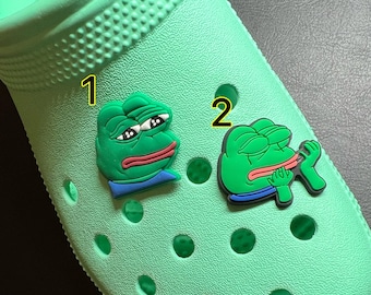 May include: Two green frog-themed shoe charms, one with a sad expression and the other with a tongue sticking out, attached to a pair of green Crocs.