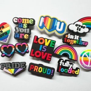 May include: A set of 11 colorful shoe charms with rainbow designs and messages of love, pride, and inclusivity. The charms feature phrases like "Love is Love", "Come as you are", "Over it", "In it together", "Fully loaded", and "Proud!"