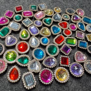 May include: A collection of colorful rhinestone embellishments in various shapes, including round, oval, square, and teardrop. The gemstones are in shades of red, pink, green, blue, yellow, and purple, each with a silver-colored border.