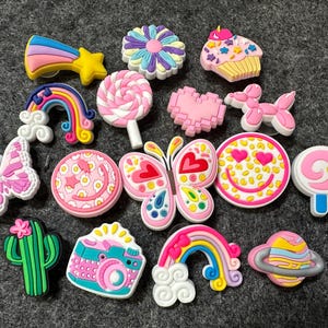 May include: A collection of 14 colorful, whimsical shoe charms featuring various designs, including a rainbow, a butterfly, a lollipop, a flower, a cupcake, a smiley face, a cactus, a camera, a star, and a balloon.