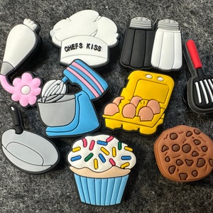 May include: A set of ten colorful silicone charms featuring kitchen-themed designs. The charms include a chef's hat, a whisk, a spatula, a cookie, a cupcake, a piping bag, a pan, a carton of eggs, and two salt shakers. The charms are all in bright colors and have a fun, whimsical design.