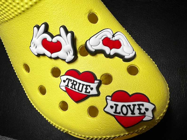 May include: Four colorful heart-shaped shoe charms with black outlines. Two charms feature a pair of hands forming a heart shape. One charm features a red heart with a black banner that says "TRUE". The last charm features a red heart with a black banner that says "LOVE".