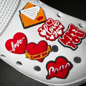 May include: White clogs with red and yellow heart-shaped charms. The charms have different messages, including "I love you", "love", and "miss you".