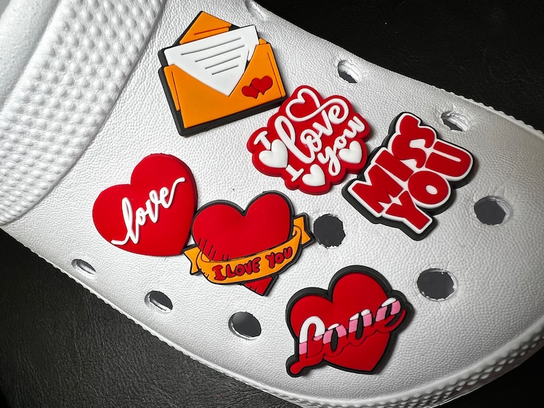 May include: White clogs with red and yellow heart-shaped charms. The charms have different messages, including "I love you", "love", and "miss you".