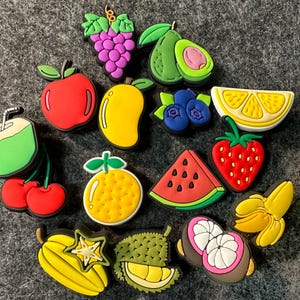 May include: A set of 13 colorful fruit and vegetable charms, including a red apple, a green coconut, a yellow mango, a yellow starfruit, a green avocado, a red watermelon, a yellow lemon, a red strawberry, a yellow orange, a green durian, a purple bunch of grapes, a yellow banana, and a pair of blue blueberries.