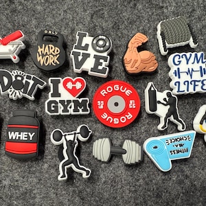 May include: A collection of colorful, themed shoe charms featuring fitness and gym-related designs. The charms include a dumbbell, a weight plate, a protein shaker, a gym bag, a barbell, a boxing glove, a heart with a barbell, a weightlifting belt, and a gym logo with the words "Gym Life".