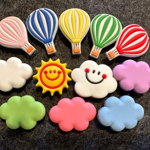 May include: A set of colorful hot air balloon and cloud charms with a yellow sun charm. The charms are made of silicone and have a smooth, glossy finish.