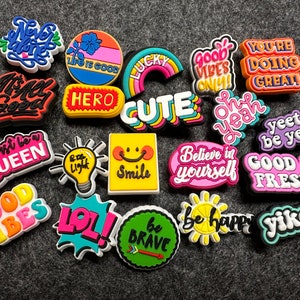 May include: A collection of colorful,  brightly colored,  plastic charms with positive messages.  The charms feature phrases like "Good Vibes Only", "Be Brave", "Just Be A Queen", "It's All Good", "Hero", "Cute", "Smile", "Believe In Yourself", "Good & Fresh", "Yeet Or Be Yeeted", "You're Doing Great", "Oh Yeah", "Yike's", "Be Happy", "Lol", "Never Alone", "Life Is Good", "Lucky", and "Good Vibes".
