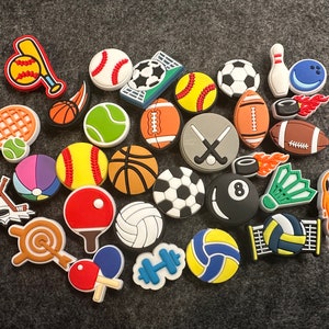 May include: A collection of colorful sports-themed shoe charms featuring various sports equipment and balls, including baseballs, basketballs, soccer balls, tennis balls, volleyballs, a badminton shuttlecock, a hockey stick, a tennis racket, a golf club, a bowling pin, a bowling ball, a football, and a basketball hoop.