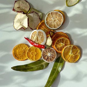 Puede incluir: Rebanadas de fruta seca y hojas dispuestas sobre una superficie blanca. La fruta incluye rodajas de naranja, limón, manzana y lima. También hay un chile rojo.