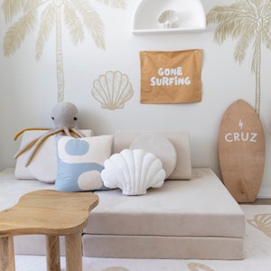 May include: A white sofa with a light blue and white patterned cushion, a white seashell cushion, and a grey octopus plush toy. The sofa is in front of a white wall with palm tree and seashell decals. A wooden surfboard with the text "CRUZ" is leaning against the wall. A wooden table with a unique shape sits in front of the sofa. The floor is covered in a white rug with a brown pattern.