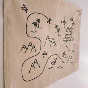May include: Tan fabric wall hanging with a black illustrated map design. The map includes mountains, palm trees, a ship, a compass, and a whale. The fabric has metal grommets in the corners for hanging.