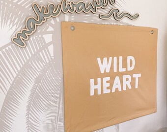 May include: A tan fabric banner with the words "WILD HEART" in white block letters. Above the banner, the words "make waves" are displayed in a cursive font, with a gray outline and a wooden base. The background features a palm tree pattern.