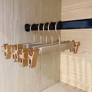 Pode incluir: Um conjunto de cabides de madeira com clipes de metal dourado. Os cabides estão pendurados em uma barra de metal preta em um armário.