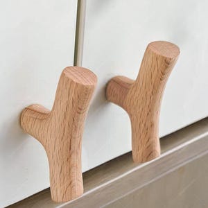 May include: Two wooden branch-shaped wall hooks. The hooks are made of light-coloured wood and have a natural grain pattern. They are mounted on a white surface, offering a rustic and minimalist design for home organisation.