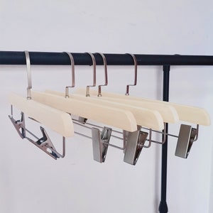 Pode incluir: Um conjunto de cinco cabides de madeira para calças com clipes e ganchos de metal prateado. Os cabides são feitos de madeira clara e estão pendurados numa barra de metal preta. Os clipes são projetados para segurar as calças com segurança.