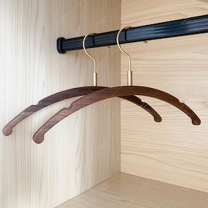 May include: Two wooden clothes hangers with brass hooks hang from a black rod inside a wooden closet. The hangers are a dark brown color and have a curved design. The closet interior is light-colored wood.