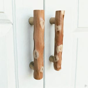 Peut inclure: Deux poignées de placard verticales fabriquées en bois naturel brun clair, présentant une forme cylindrique avec des nœuds visibles et une surface texturée rappelant l'écorce. Chaque poignée est fixée sur une porte blanche à panneaux.