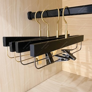 Op de afbeelding: Set van drie zwarte houten hangers met gouden metalen haken en clips. De hangers hangen aan een zwarte metalen stang.