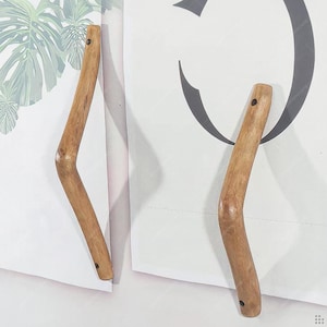 May include: Two wooden coat hooks with a curved design. The hooks are made of light brown wood and have a natural, organic look. Each hook has a small hole at the top and bottom for mounting. The hooks are mounted on a white background.