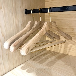 Puede incluir: Tres perchas de madera con ganchos dorados colgadas de una barra negra dentro de un armario de madera. Las perchas son de color marrón claro con un acabado de veta de madera natural. El interior del armario también es de madera clara.