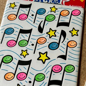 Puede incluir: Una hoja de pegatinas con más de 125 caras sonrientes de colores con notas musicales. Las pegatinas están sobre un fondo blanco con líneas onduladas azules.