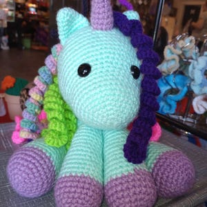 Puede incluir: Un juguete de unicornio de crochet con un cuerpo azul claro, una melena y una cola moradas, y detalles verdes y rosas. El unicornio tiene un cuerno morado y ojos negros.