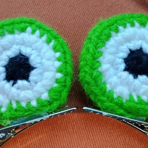 May include: Two green and white crocheted eye-shaped hair clips with black centers. The clips are attached to silver metal alligator clips.