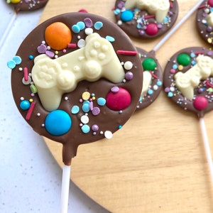 May include: A chocolate lollipop with a white chocolate video game controller design. The lollipop is decorated with colourful sprinkles and candy.
