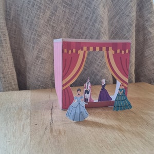 May include: A papercraft stage with red curtains and four figures in costumes. The figures are a king, a queen, and two women in ball gowns. The stage is made of cardboard and has a cutout window.