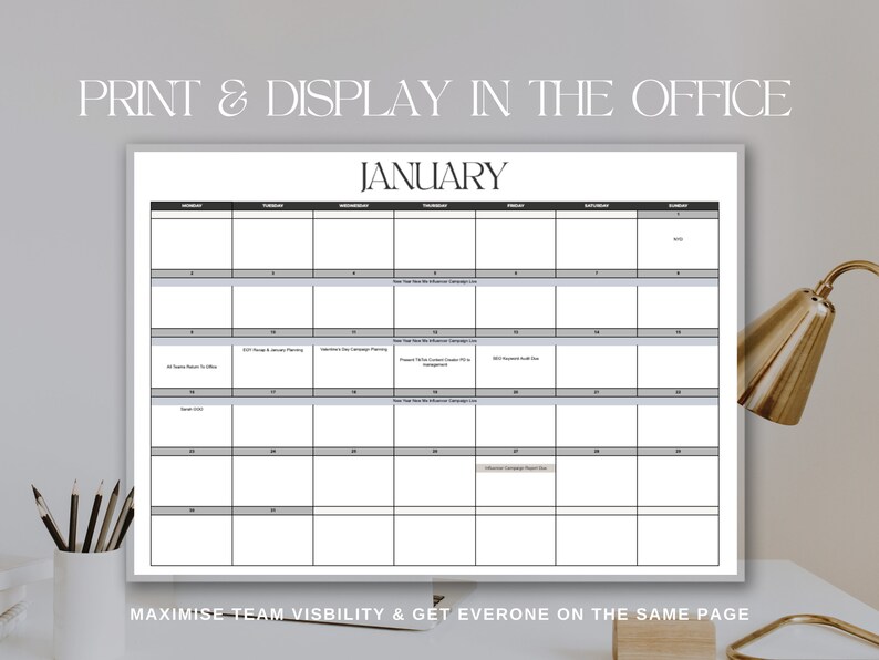 A mock up of the January month calendar hanging on a wall is showed infront of a background of a desk in an office setting with pencils in a caddy and a gold lamp. Text reads: Print & Display In The Office, Maximise team visibility