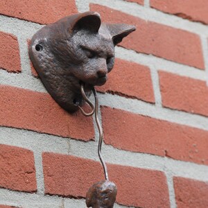 May include: A brown cast iron cat head door knocker with a metal tongue hanging down. The knocker is mounted on a brick wall.