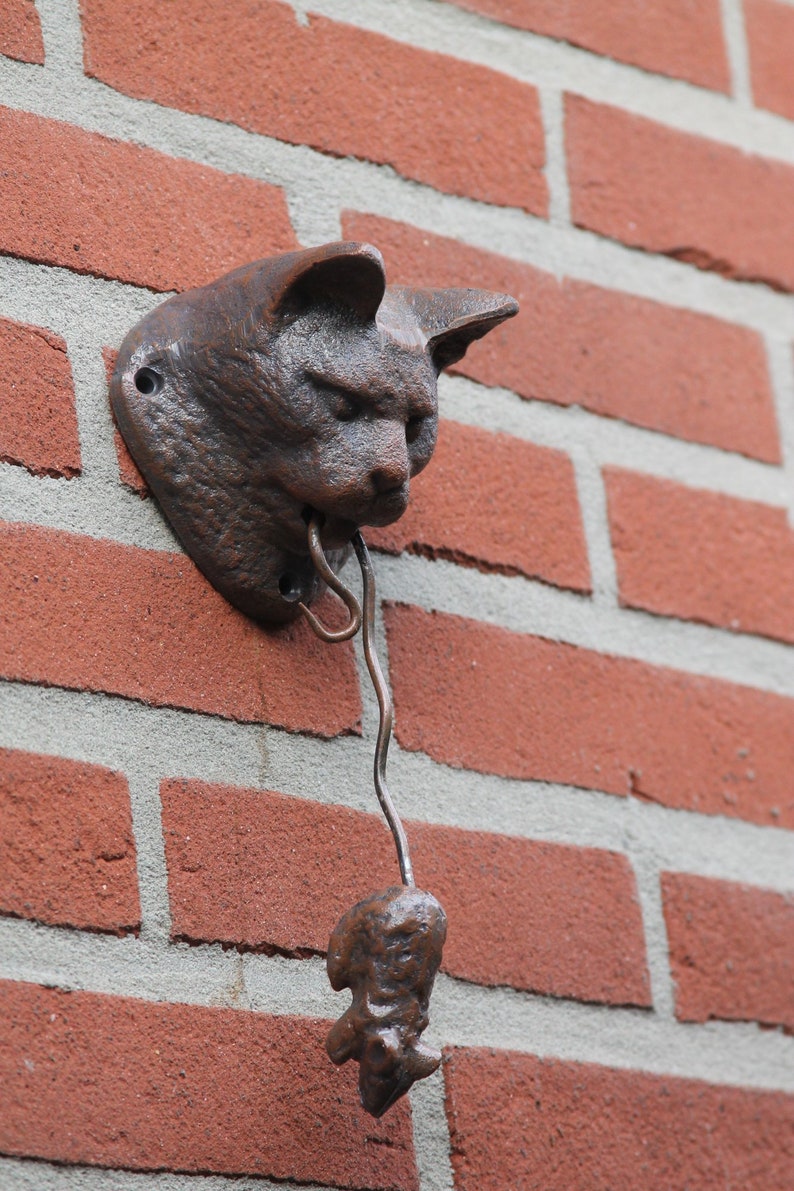 May include: A brown cast iron cat head door knocker with a metal tongue hanging down. The knocker is mounted on a brick wall.