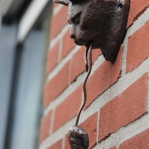 May include: A brown metal cat figurine with a mouse in its mouth. The cat is mounted on a brick wall.