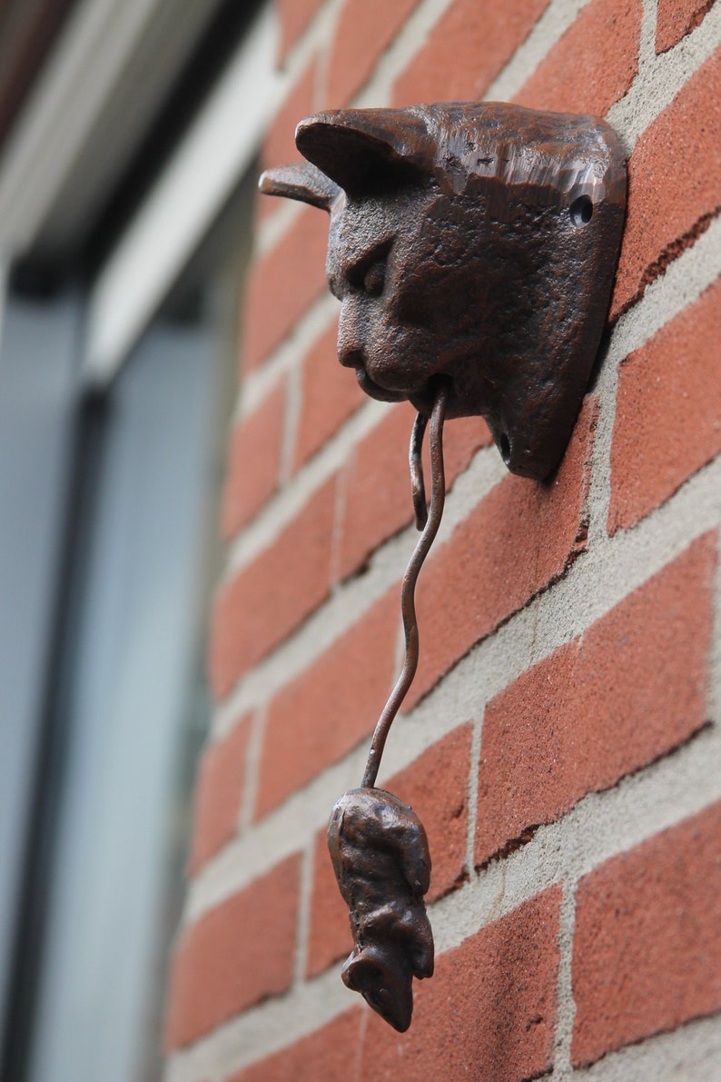 May include: A brown metal cat figurine with a mouse in its mouth. The cat is mounted on a brick wall.