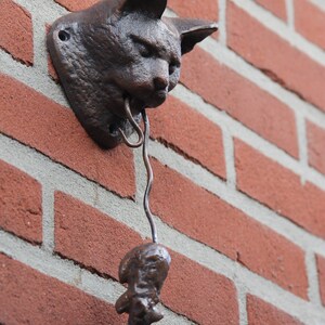 May include: A cast iron cat head door knocker with a hanging bell. The cat head is brown and has a realistic design. The bell is attached to the cat's mouth by a metal chain.