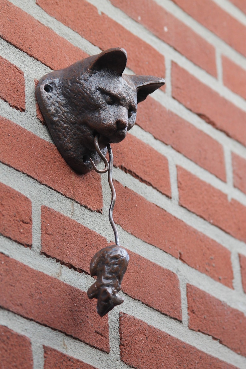 May include: A cast iron cat head door knocker with a hanging bell. The cat head is brown and has a realistic design. The bell is attached to the cat's mouth by a metal chain.