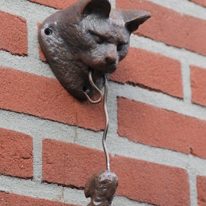 May include: A brown cast iron cat door knocker with a mouse hanging from its mouth. The knocker is mounted on a brick wall.