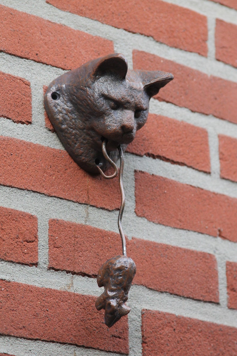 May include: A brown cast iron cat door knocker with a mouse hanging from its mouth. The knocker is mounted on a brick wall.