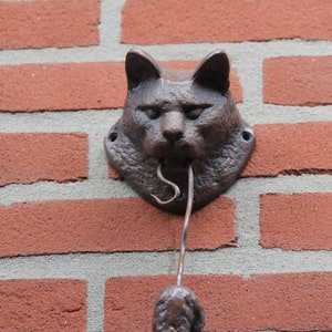 May include: A brown metal cat head door knocker with a mouse-shaped pull handle attached to a brick wall.