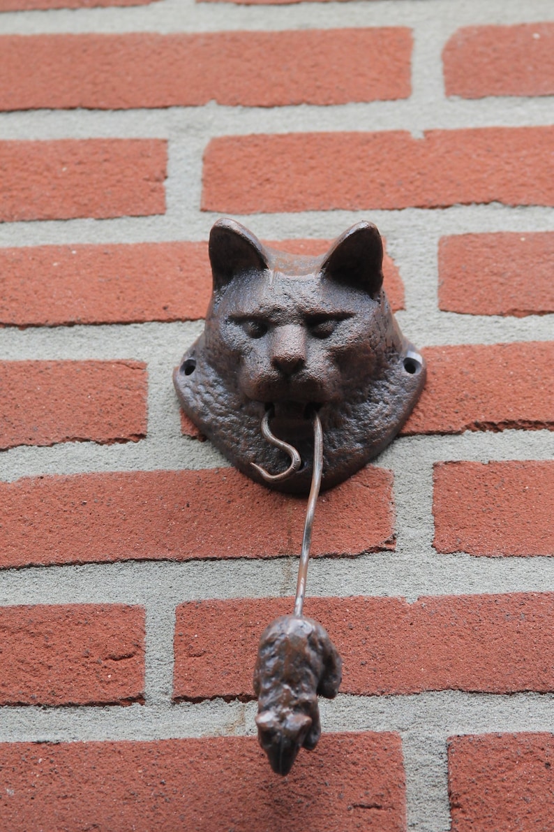 May include: A brown metal cat head door knocker with a mouse-shaped pull handle attached to a brick wall.