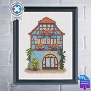 Peut inclure: Modèle de point de croix d'une maison bleue et blanche avec un toit rouge et des finitions brunes. La maison a une grande porte cintrée et des fenêtres avec des jardinières. Il y a des plantes vertes et des arbres autour de la maison.