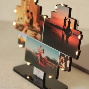 May include: A heart-shaped photo frame with a black wooden base and warm white LED lights. The frame is made up of multiple smaller frames, each displaying a different photograph of a couple. The photos show the couple in various settings, including a beach, a city, and a park.