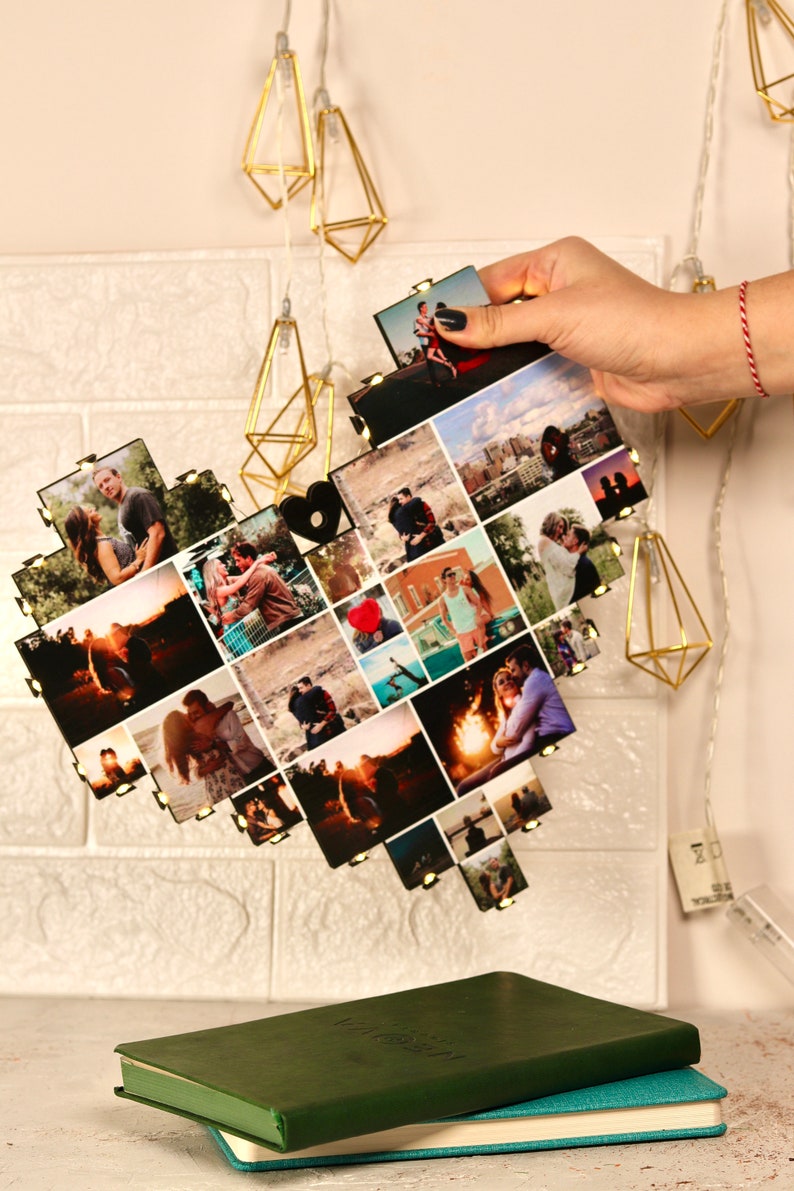 May include: A heart-shaped photo collage with lights, featuring a couple's photos. The collage is made of wood and has a black finish. The photos are arranged in a grid pattern and show the couple in various settings, including a campfire, a city street, and a beach.