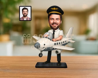 以下が含まれることがあります： 白とグレーの飛行機に乗ったパイロットの気まぐれな置物。 パイロットは黒い帽子と制服を着ています。 背景には男性の小さな額入り写真があります。 置物は黒い台座の上にあります。