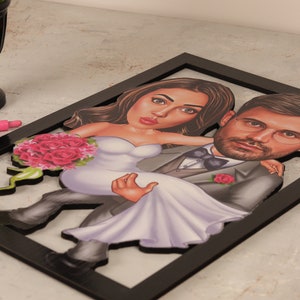 May include: A black framed 3D caricature of a couple in wedding attire. The groom is holding the bride in his arms. The bride is wearing a white dress and a bouquet of pink roses. The groom is wearing a gray suit with a black bow tie and a pink rose on his lapel.