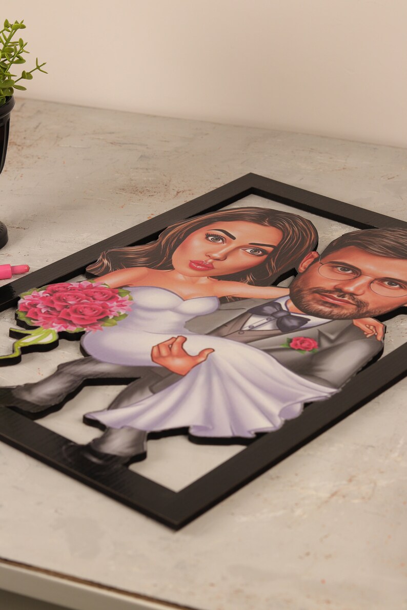 May include: A black framed 3D caricature of a couple in wedding attire. The groom is holding the bride in his arms. The bride is wearing a white dress and a bouquet of pink roses. The groom is wearing a gray suit with a black bow tie and a pink rose on his lapel.