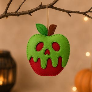 May include: A felt apple ornament featuring a red base with a bright green dripping glaze overlay. The green section includes cutouts for two oval eyes and a small heart-shaped nose, creating a skull-like appearance. A brown stem and green leaf are attached to the top with a thin hanging string.