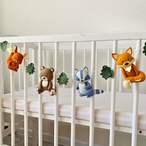 Op de afbeelding: Een vilten slinger met bosdieren met een eekhoorn, beer, wasbeer en vos, geregen met groene vilten bladeren. De slinger wordt weergegeven op een wit ledikant, waardoor een grillige kinderkamerdecoratie ontstaat.